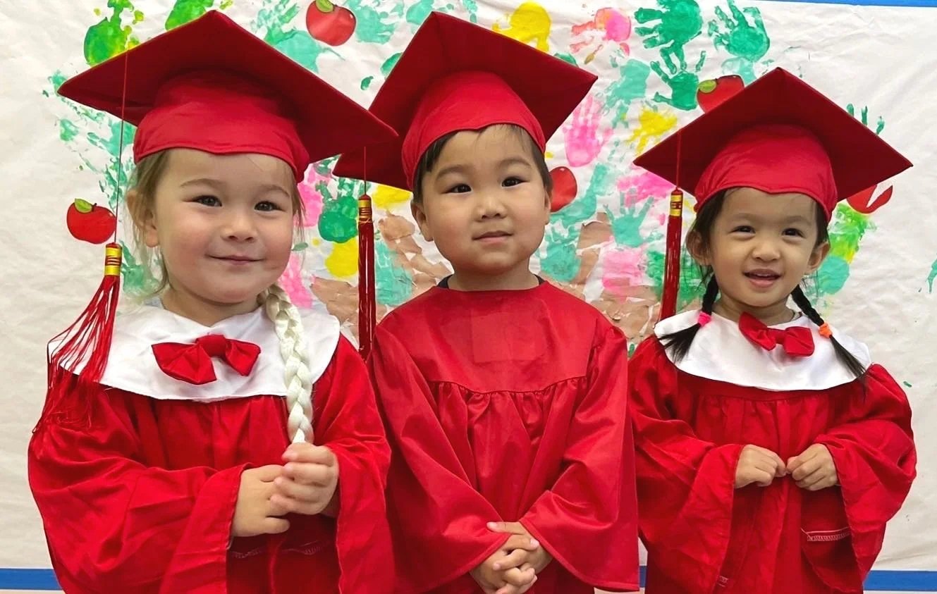 Three Apple Tree preschoolers in red graduation caps and gowns