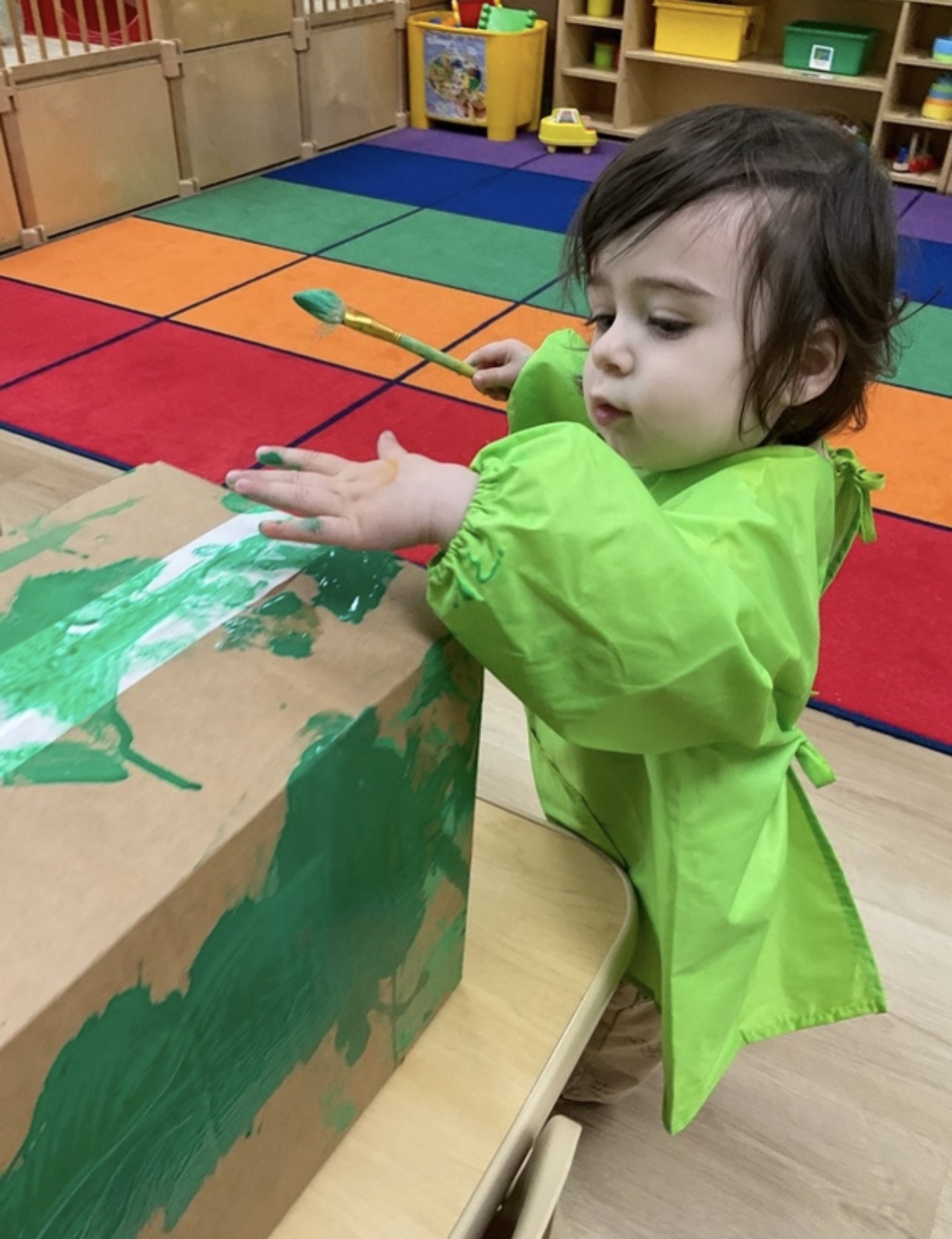 An Apple Tree toddler in a green smock painting a large cardboard box with green paint