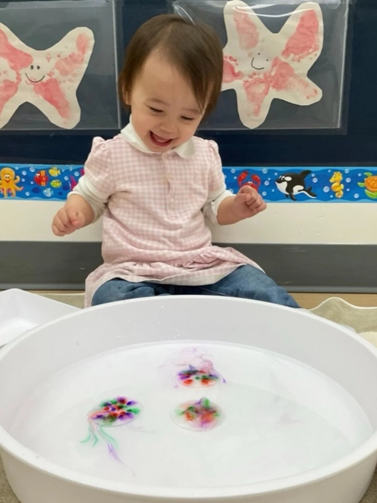An Apple Tree infant laughing beside a shallow water tray during a sensory activity